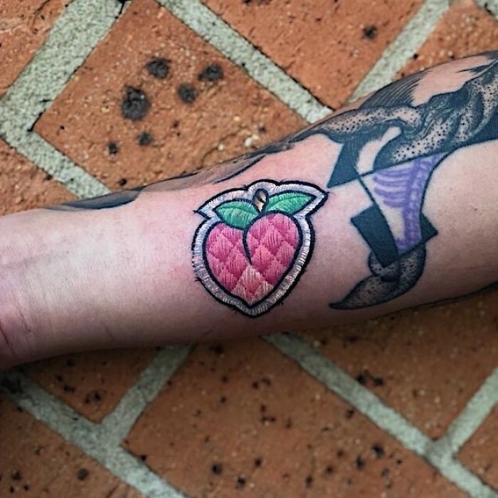 Tattoo resembling a sewn-on patch with a peach design on a person's forearm.