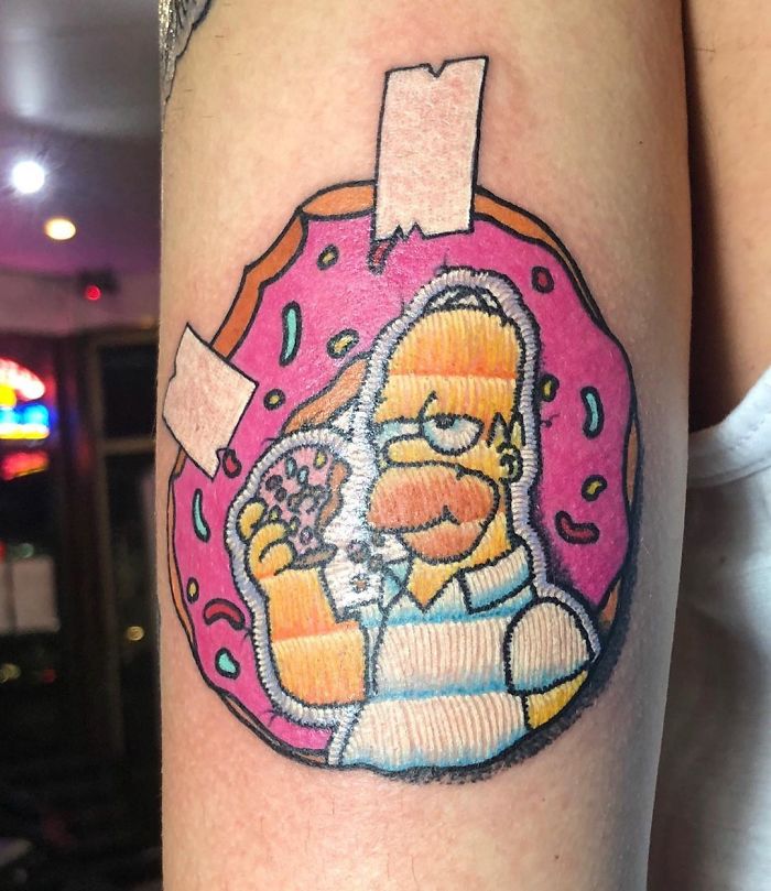 Tattoo resembling a sewn-on patch of a cartoon character with a pink donut, featuring a unique embroidery style.
