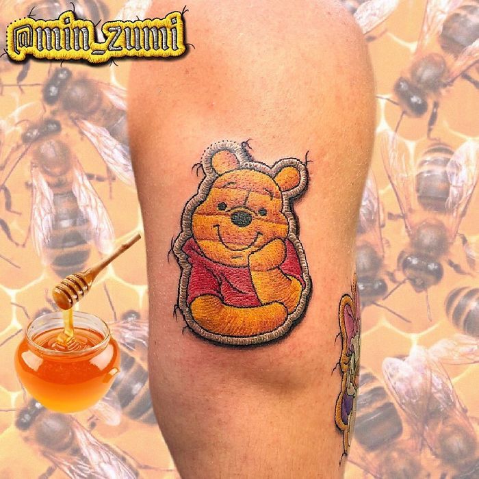 Tattoo resembling a sewn-on patch of a bear in a red shirt on an arm, honey and bees in the background.