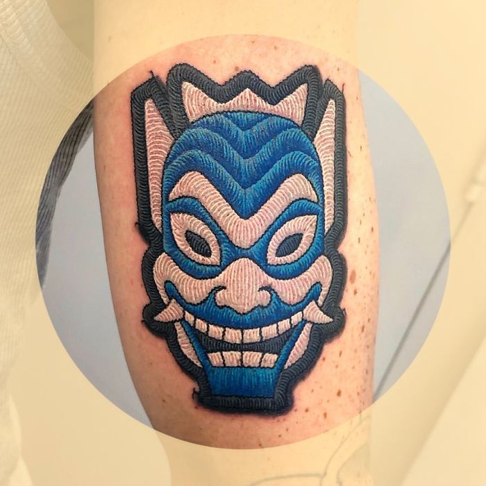 Tattoo resembling a blue and pink sewn-on patch on an arm, showcasing intricate details and texture.