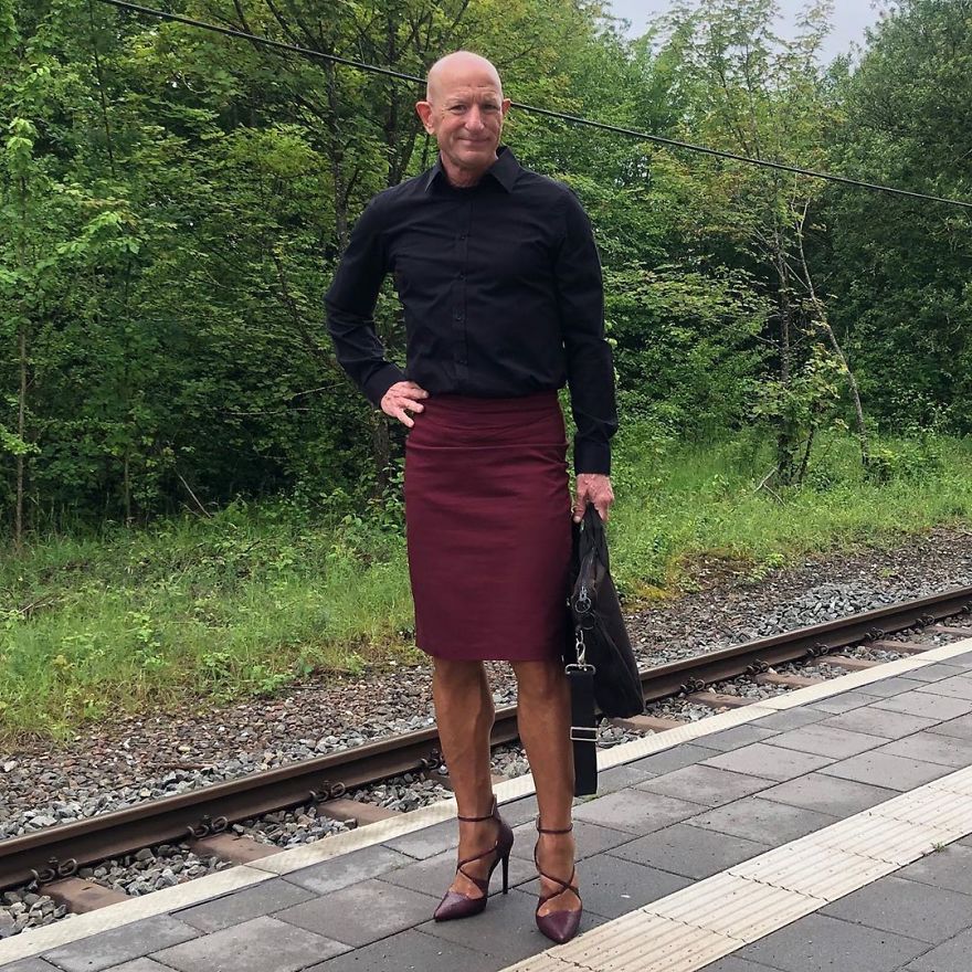 Mark Bryan is wearing a black shirt, bordo skirt and high heels and holding a briefcase