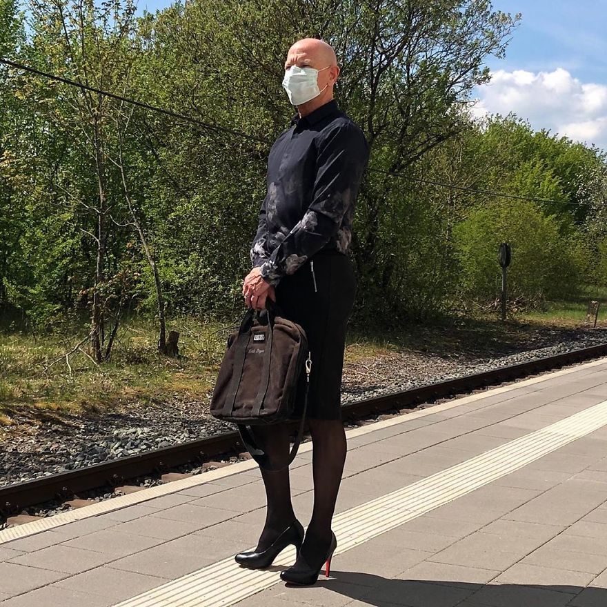 Mark Bryan is standing outside near the railway in a black outfit with a shirt, skirt, and high heels and holding a briefcase