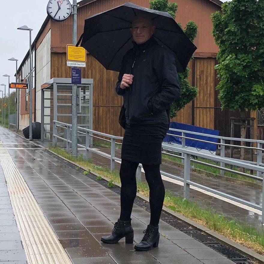 Mark Bryan is standing in the rain in a black outfit with a jacket, skirt, and high heel boots and holding an umbrella