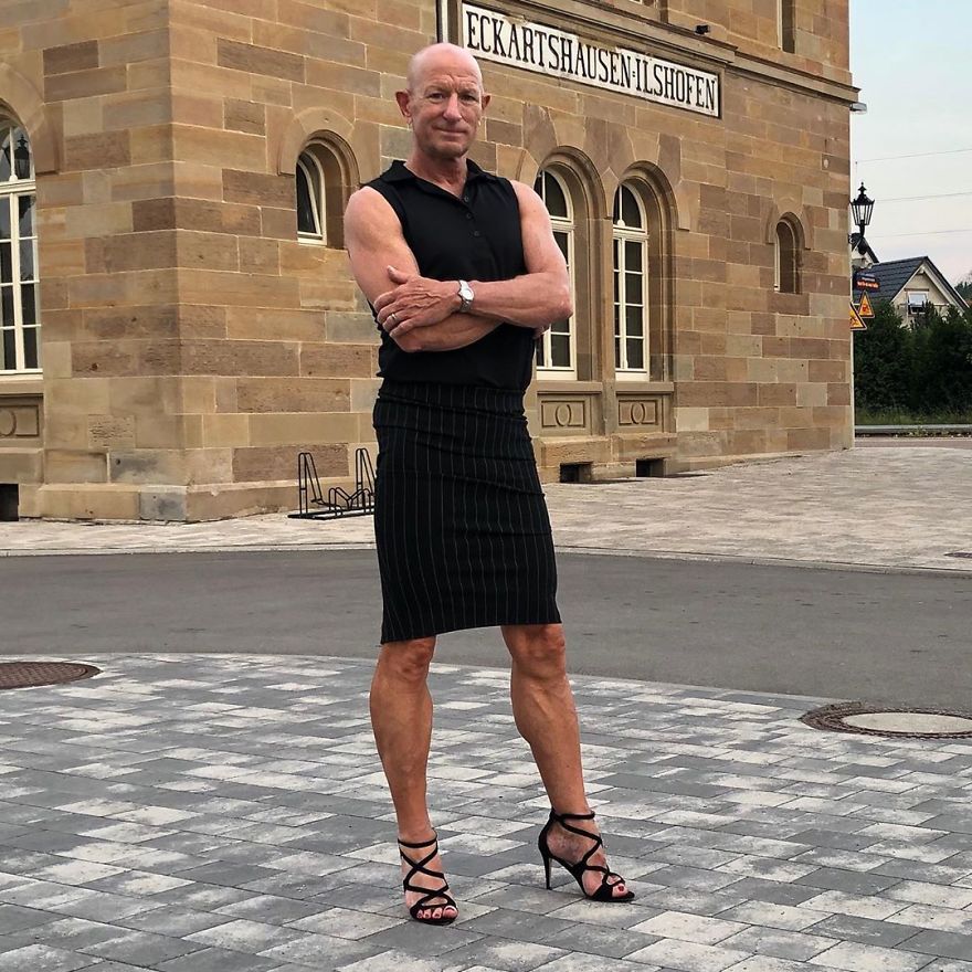 Mark Bryan standing outside in a black outfit with a sleeveless shirt, striped skirt, and high heels