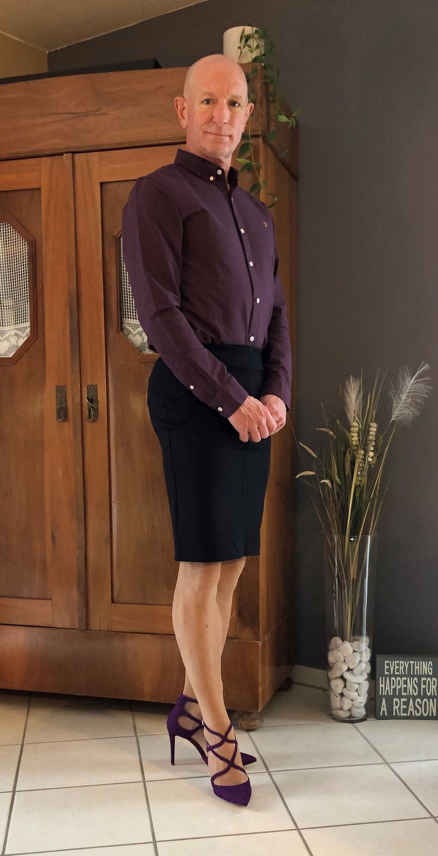Mark Bryan is wearing an aubergine color shirt and high heels, a black skirt and standing in front of a wooden wardrobe