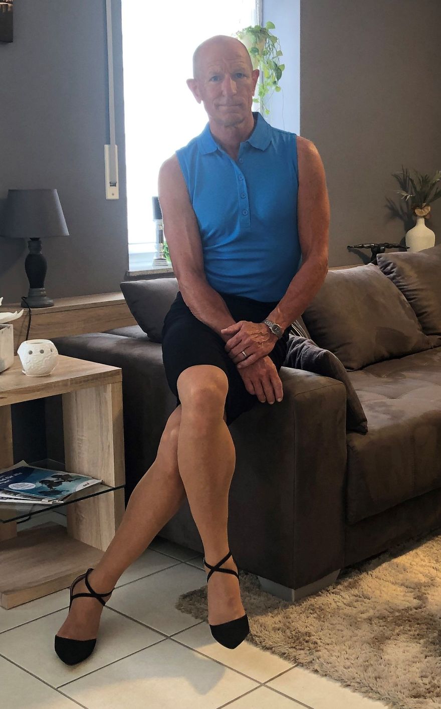 Mark Bryan is wearing a blue sleeveless shirt, black skirt, and high heels and sitting on the edge of the sofa
