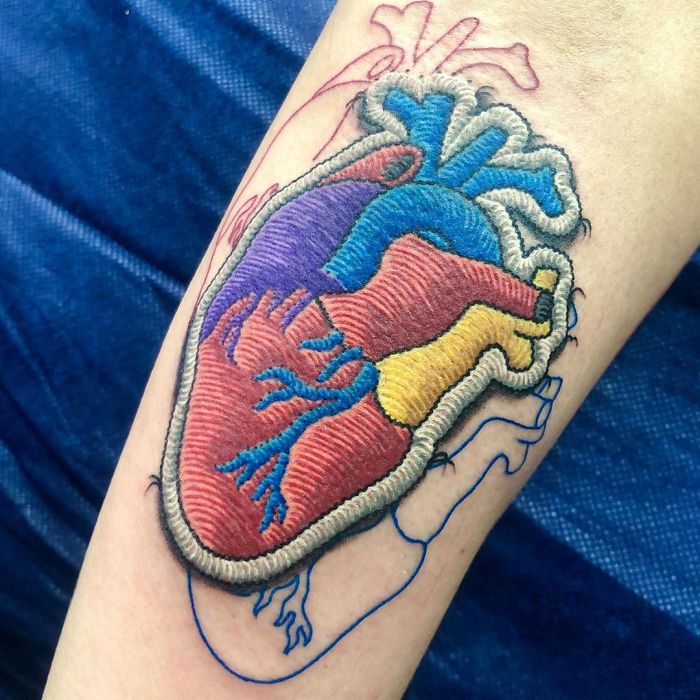 Tattoo resembling a colorful sewn-on patch of an anatomical heart on forearm.