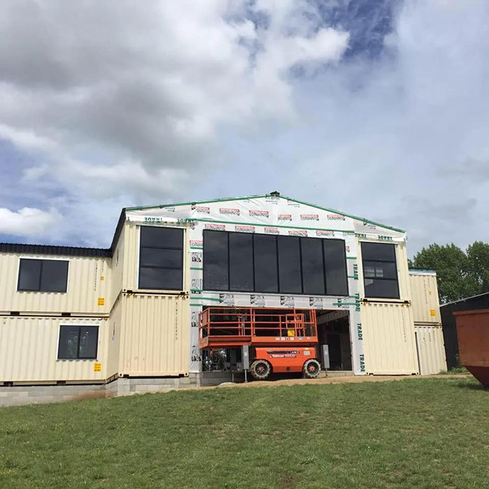 Shipping container house under construction with large windows and an orange lift in front.