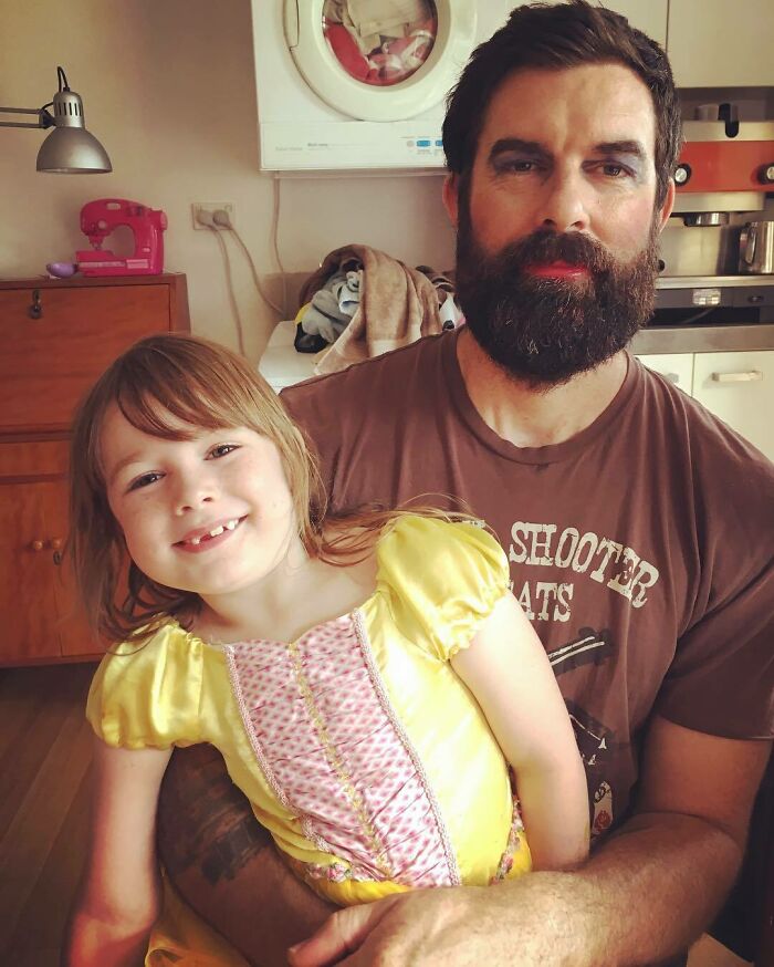 Dad with lipstick on his beard holding smiling daughter in a yellow dress after a visit to their beauty salon playtime.