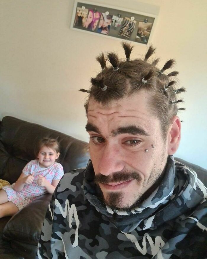 Dad with playful hairstyle after a visit to his daughters beauty salon, smiling with daughter giving thumbs up on couch.