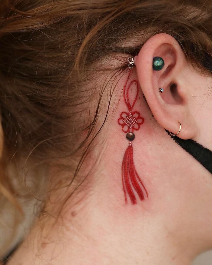 Intricate body art tattoo of a red tassel behind the ear by artist Sion Kwak.