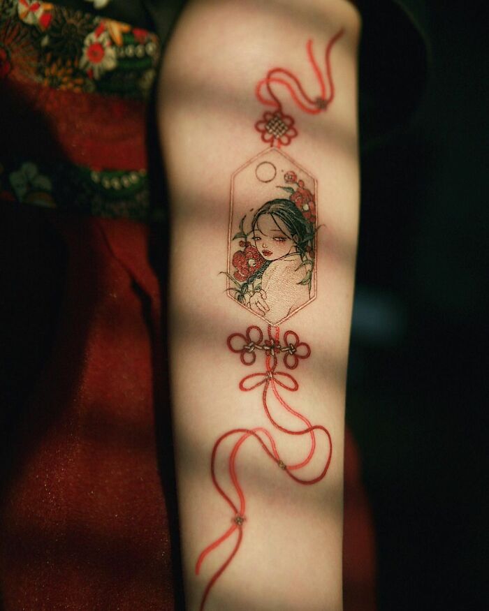 Beautiful body art with intricate design by tattoo artist Sion Kwak, featuring a delicate red ribbon and floral motif on an arm.