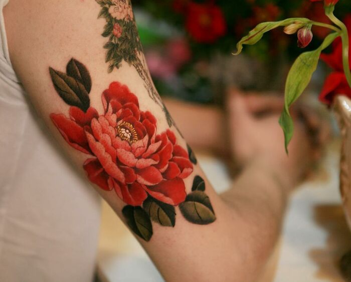 Red peony tattoo art on an arm, featuring intricate details and vibrant colors, created by artist Sion Kwak.
