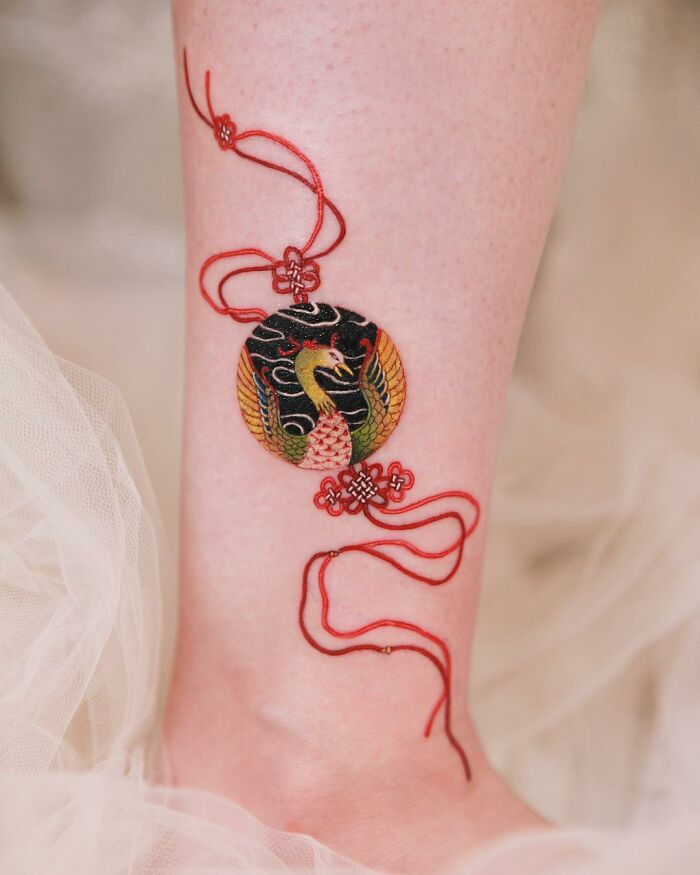 Beautiful body art tattoo by Sion Kwak featuring a colorful bird and intricate red thread design on the leg.