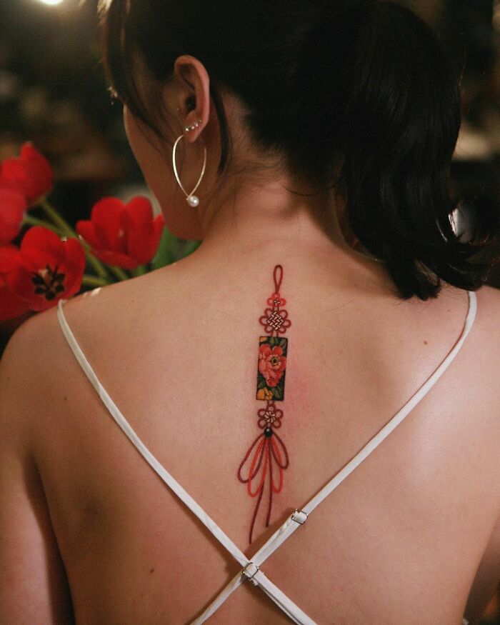 Tattoo art by Sion Kwak on a woman's back, featuring floral design with red accents, set against a backdrop of red flowers.