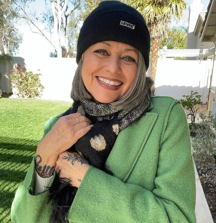 Woman in her 50s smiling outdoors, wearing a green coat, black beanie, and scarf, confidently embracing her unique style. Woman in her 50s smiling outdoors, wearing a green coat, black beanie, and scarf, confidently embracing her unique style.