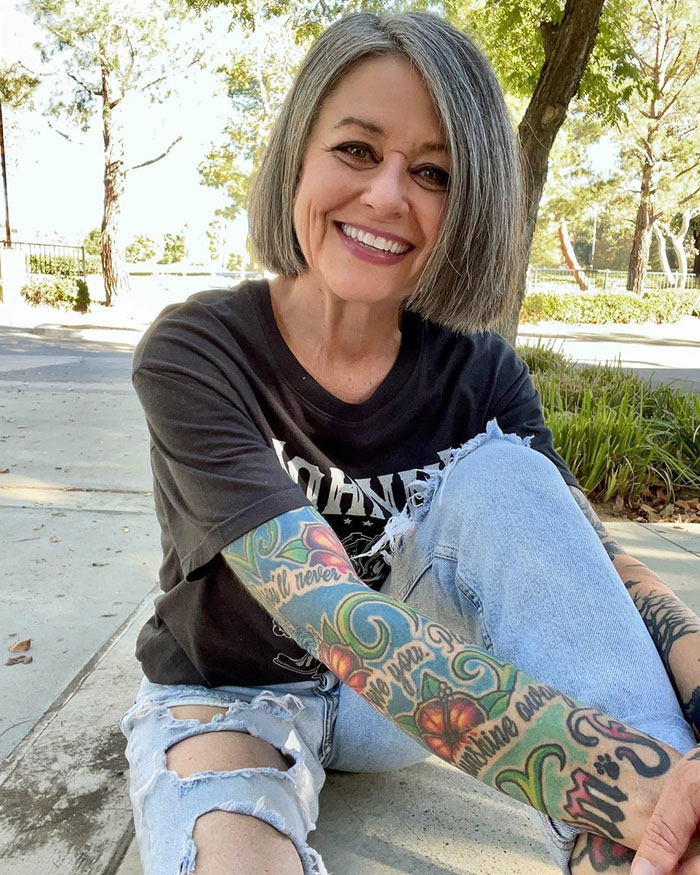 Woman in her 50s wearing a casual outfit with ripped jeans and colorful tattoos, smiling outdoors on a sunny day. Woman in her 50s wearing a casual outfit with ripped jeans and colorful tattoos, smiling outdoors on a sunny day.