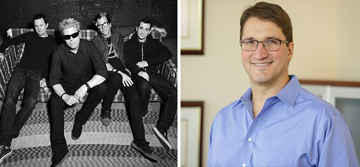 Black and white photo of a band sitting on a couch next to a man in glasses and a blue shirt smiling indoors, interesting facts.