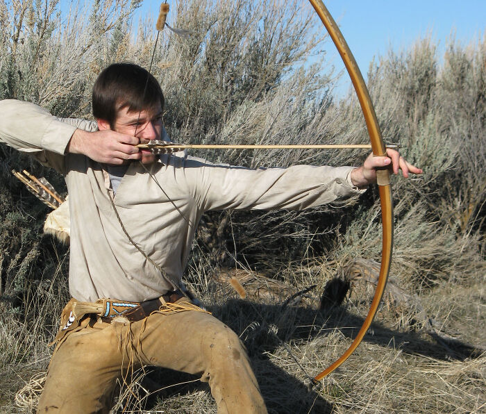 Man dressed in historical clothing practicing archery with a bow and arrow in an outdoor natural setting, interesting facts.