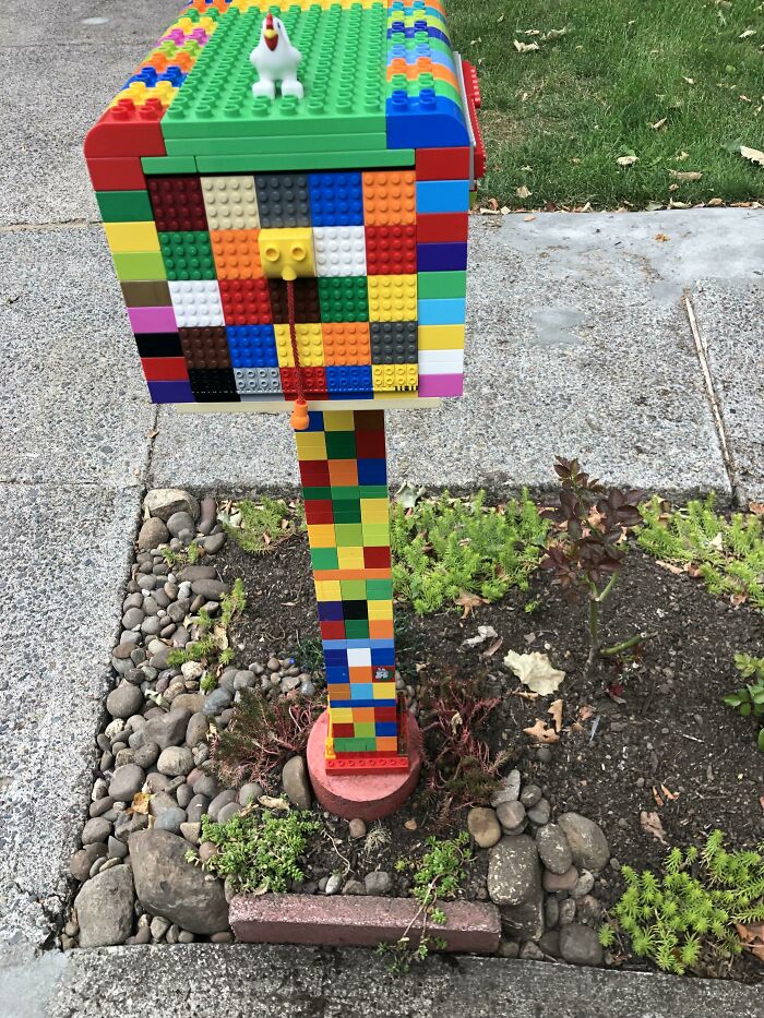Colorful creative mailbox built entirely from toy building blocks with a small rooster figure on top in a garden setting