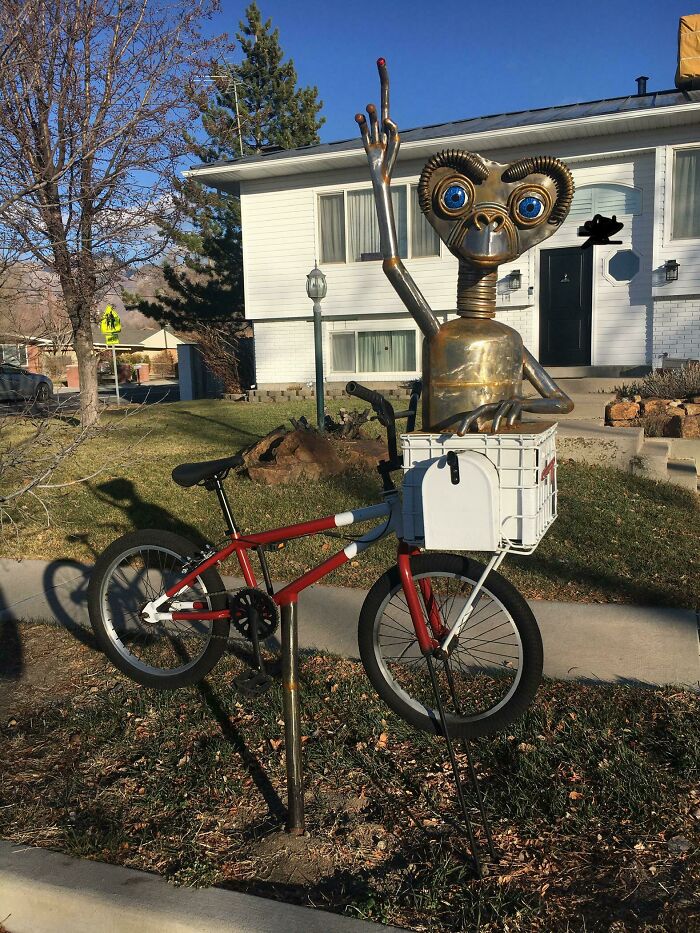 Creative and cool mailbox designed as a metal alien figure on a bike with a white mailbox attached in a residential yard.