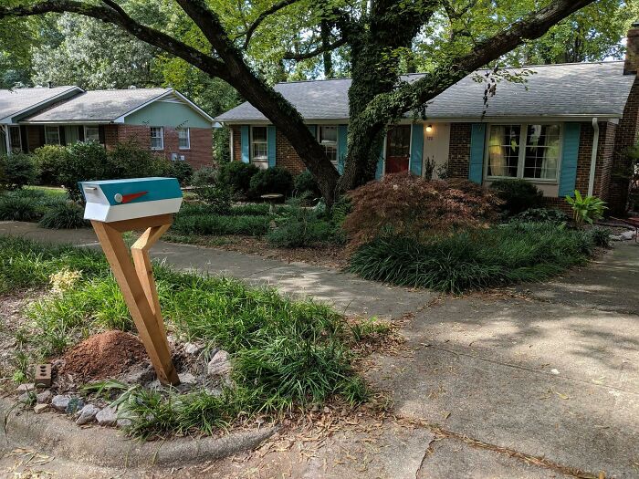 Creative and cool mailbox with a modern design stands on a wooden post near a suburban house and green landscaping.