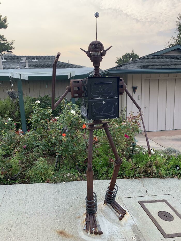 Creative and cool mailbox designed as a robot sculpture standing in a garden by a house walkway.