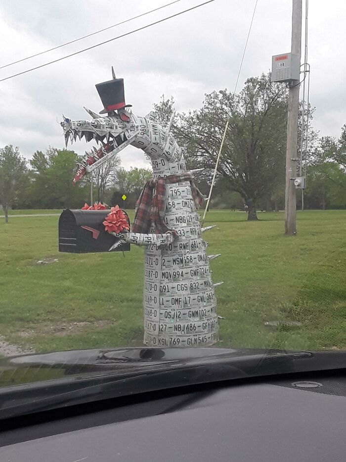 Creative mailbox made from license plates shaped like a dragon wearing a top hat and holding a black mailbox with red flowers.