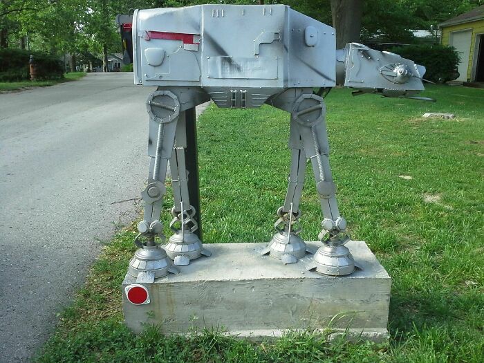 Creative and cool mailbox designed like a Star Wars AT-AT Walker, standing on a concrete base near a grassy roadside.