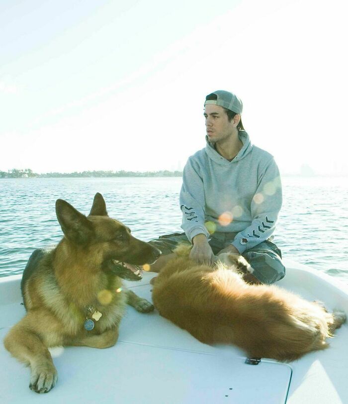 Young man with two dogs on a boat, showcasing a celebrity's love for dogs by the water on a sunny day.