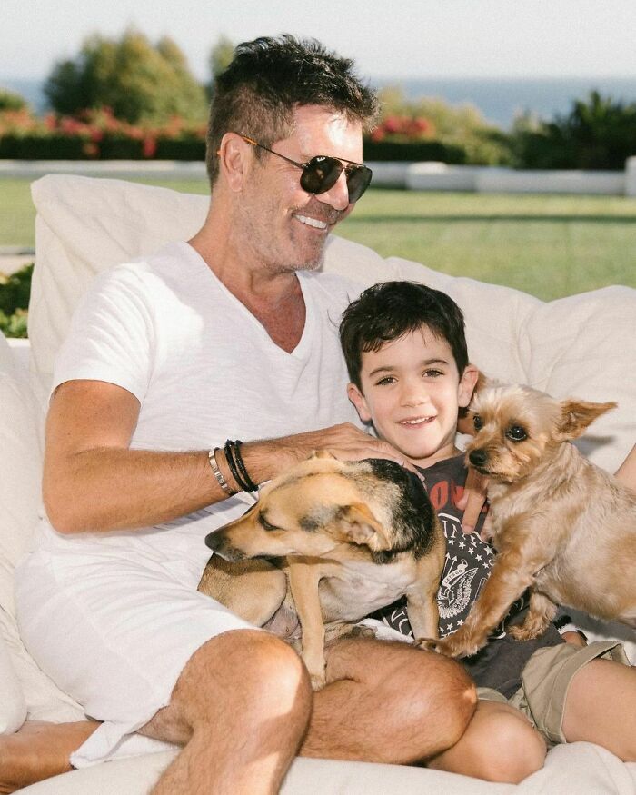 Man and child smiling outdoors, cuddling two dogs, showing immense love for dogs in a relaxed and happy moment.