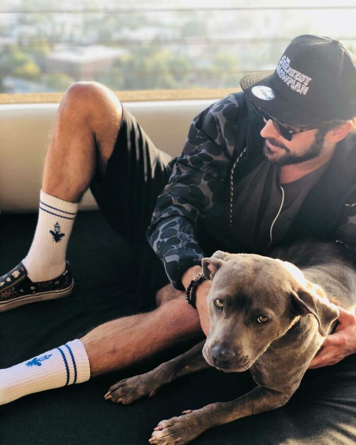 Man wearing sunglasses and cap relaxing with a gray dog, showing a celebrity's love for dogs in a casual setting.