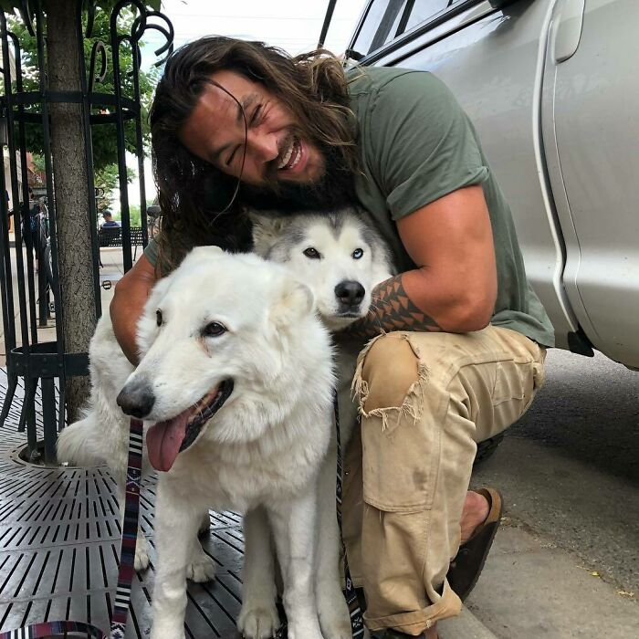 Man crouching and smiling while hugging two dogs on a city sidewalk showing love for dogs and pets outdoors.