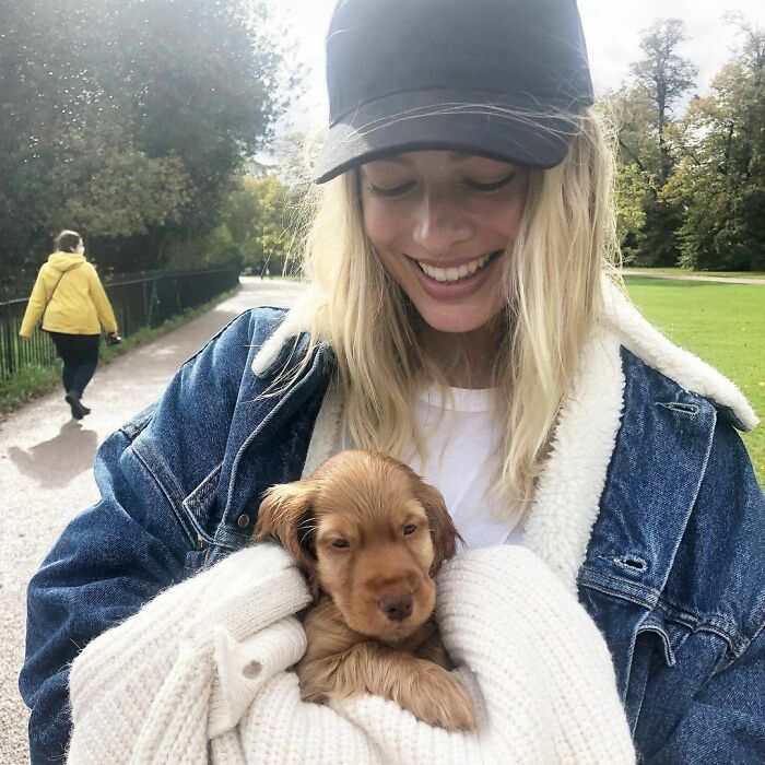 Smiling woman wearing a black cap and blue jacket holding a small brown puppy, showing immense love for dogs outdoors.
