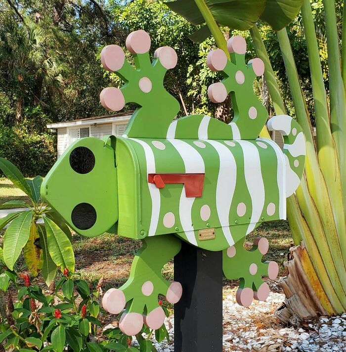 Creative and colorful mailbox designed as a green and white spotted lizard, standing outdoors among plants.