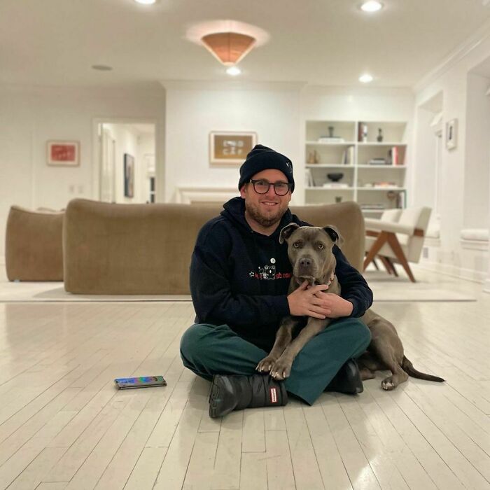 Celebrity sitting on floor of modern home, wearing casual clothes and beanie, hugging a large dog showing love for dogs.