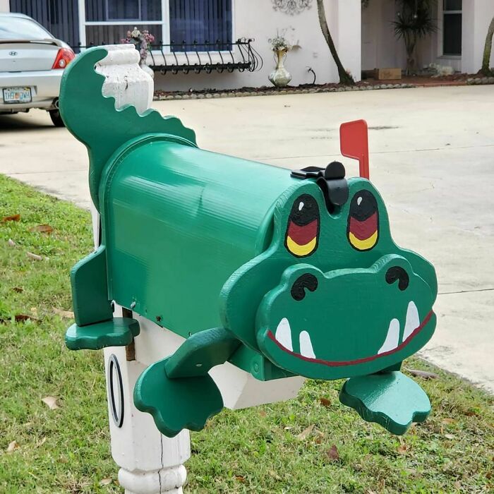 Green alligator-shaped mailbox with a red flag and colorful eyes on a white post by a suburban driveway.