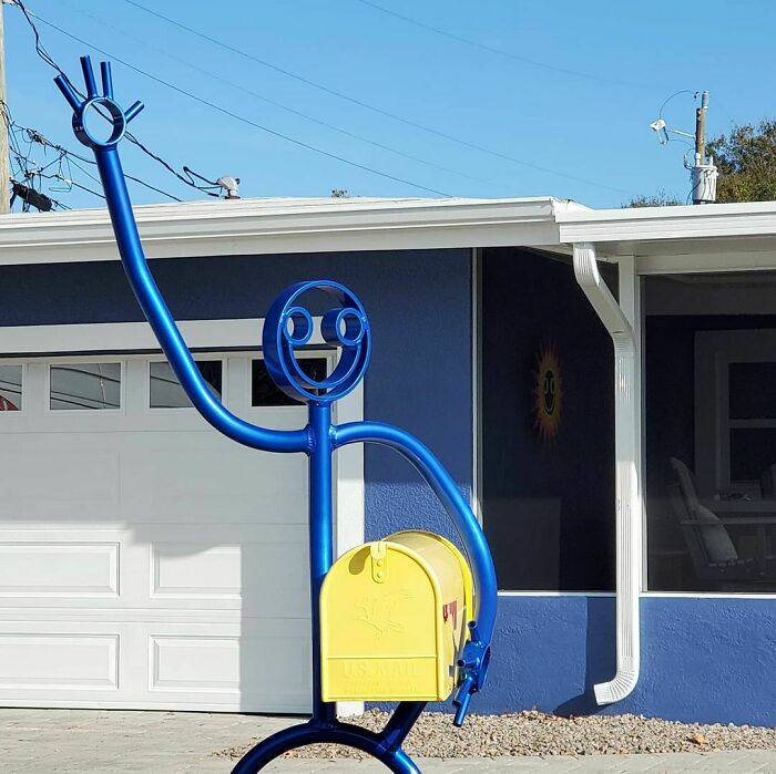 Blue metal sculpture holding a yellow mailbox with a smiling face design, showcasing creative mailboxes in the wild.