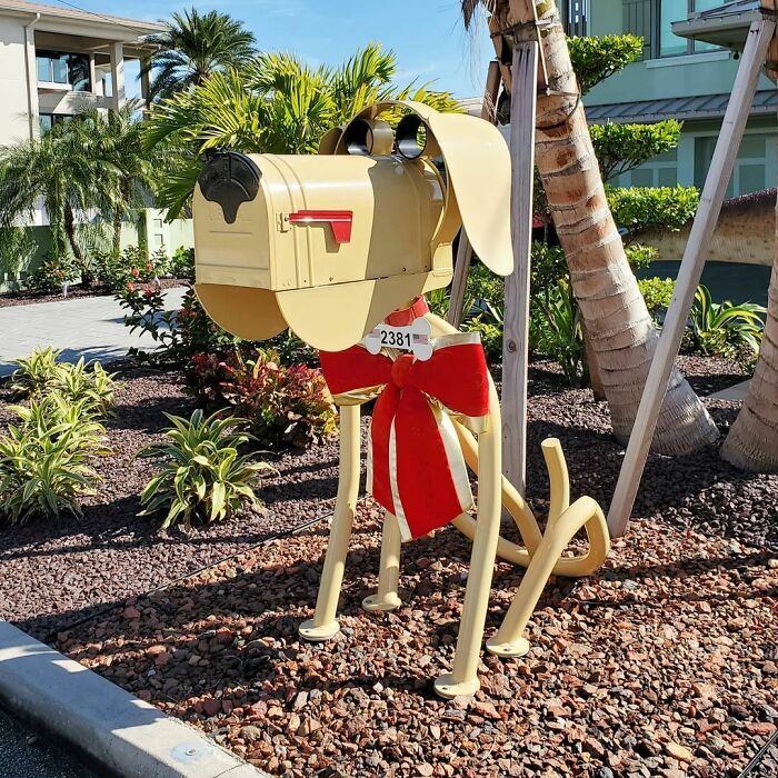 Creative and cool mailbox designed as a dog with a red bow, standing in a landscaped garden near palm trees.