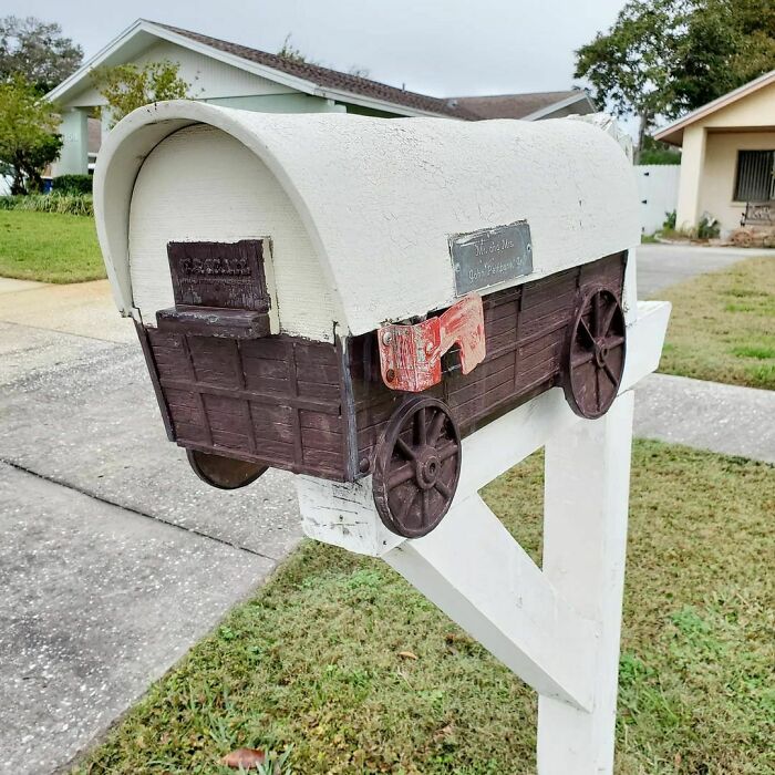 Creative and cool mailbox designed like a covered wagon with wooden wheels mounted on a white post outdoors.