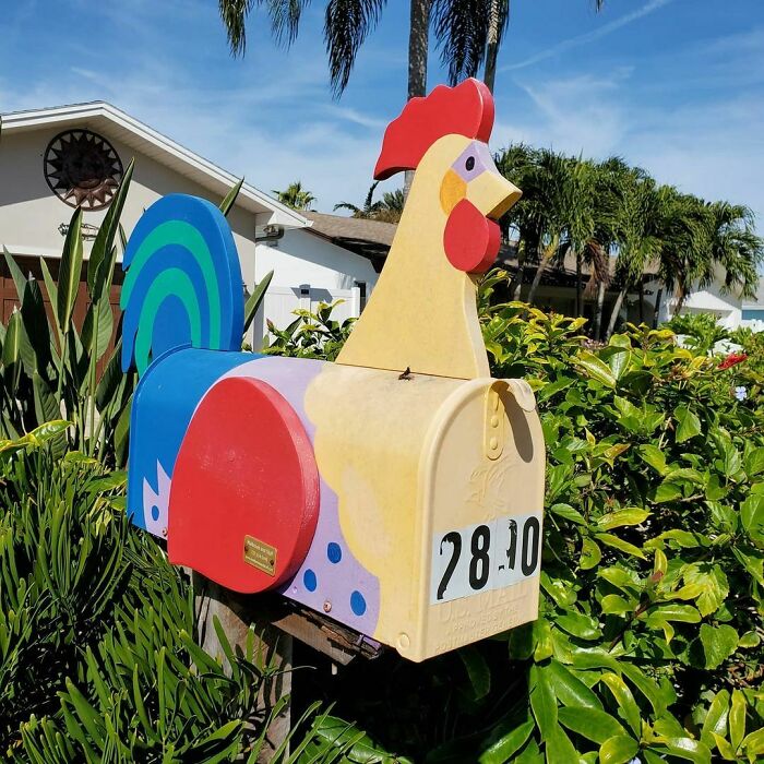 Colorful creative mailbox designed as a rooster, surrounded by greenery and set in a sunny outdoor residential area.
