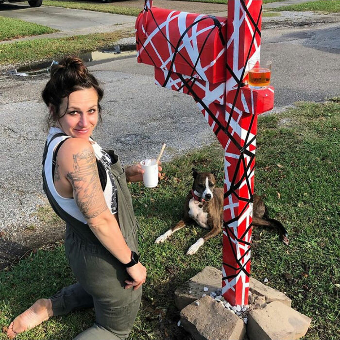 Woman painting a creative and cool mailbox with red, white, and black abstract design outdoors with her dog nearby.
