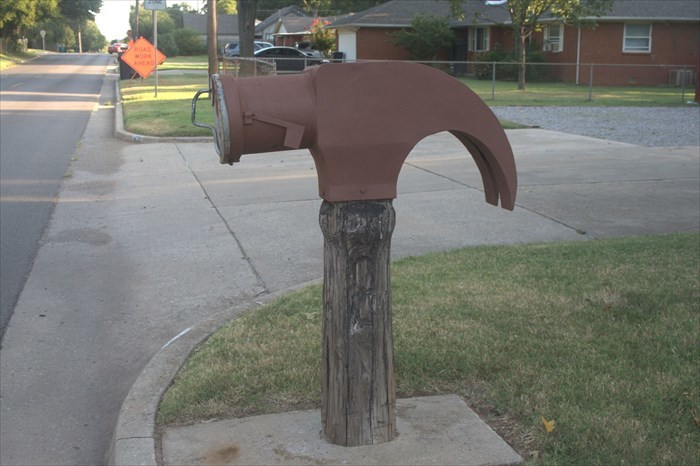 Creative and cool mailbox shaped like a large brown hammer mounted on a wooden post by a suburban street.