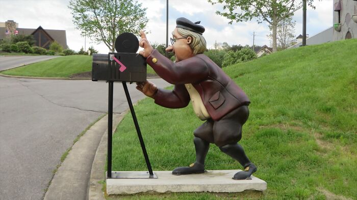Sculpture of a man opening a creative and cool mailbox designed like a vintage camera on a roadside lawn.