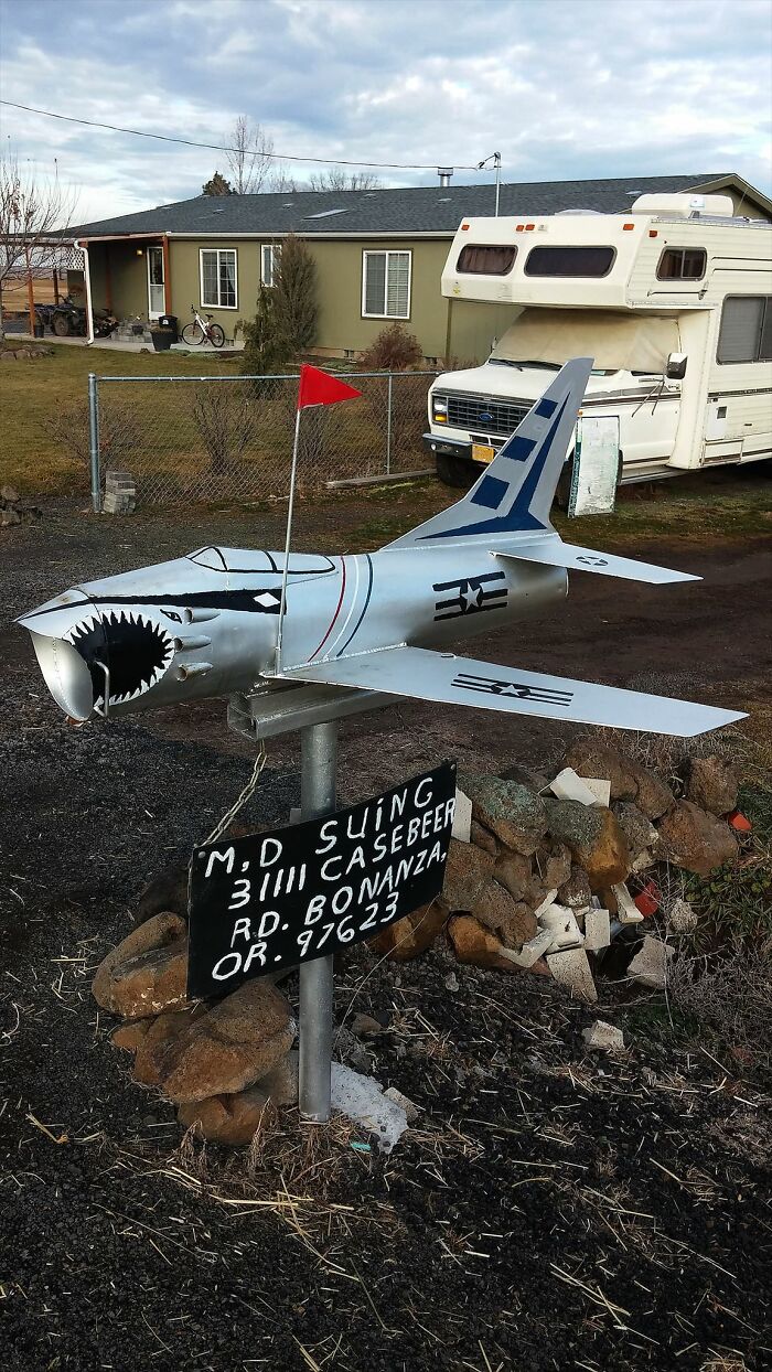 Mailbox shaped like a vintage fighter jet with shark teeth design, mounted on a pole with an address sign below.