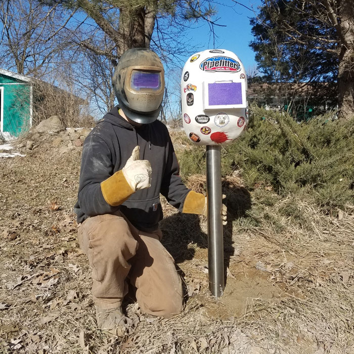 Person in welding gear giving thumbs up next to a creative and cool mailbox decorated with stickers in an outdoor setting.