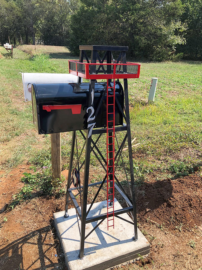 Creative and cool mailbox designed like a black tower with red ladder and railing, set in a rural outdoor area.