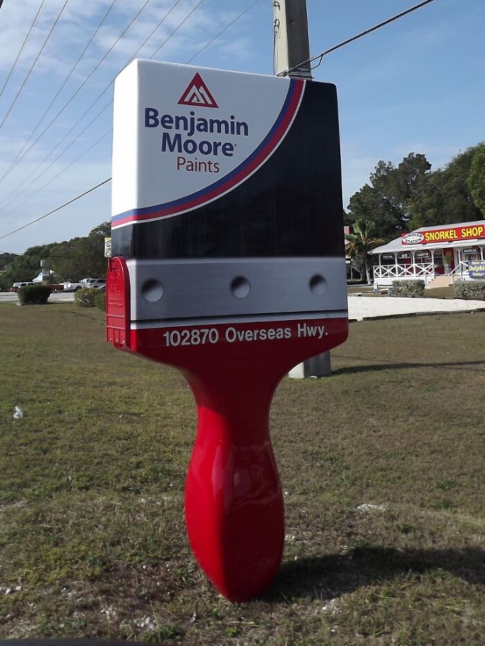 Creative mailbox shaped like a large paintbrush with Benjamin Moore branding standing on grassy roadside.