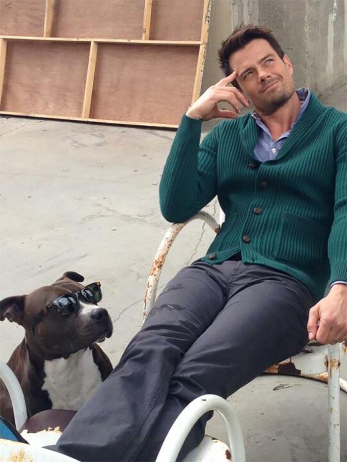 Man in a green sweater sits relaxed outdoors next to a dog wearing sunglasses showing love for dogs and celebrities.
