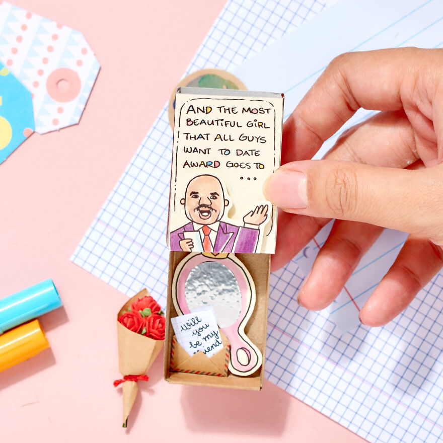 Hand holding a cute handmade card asking a girl to be your girlfriend with small paper roses and colorful pens nearby.